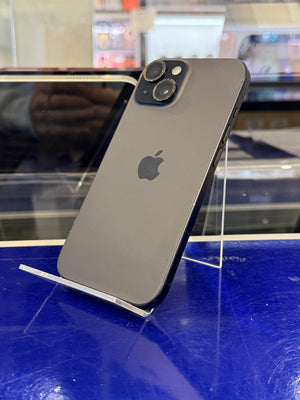 A dark gray Apple iPhone 12 Pro shown from the back, resting on a clear acrylic display stand against a bright blue wall in a retail setting.