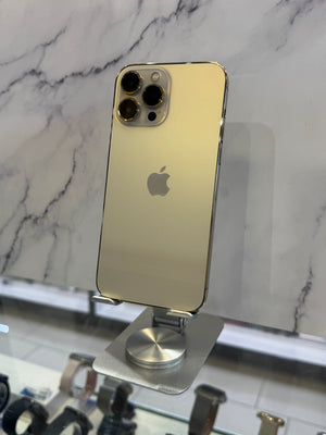 A gold-colored Apple iPhone Pro shown from the back, resting on a silver metal display stand against a white marble-patterned background.