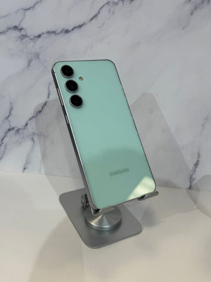A mint green Samsung smartphone shown from the back, resting on a clear stand against a white marble-patterned background.