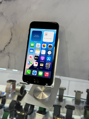 A black smartphone displayed upright on a metallic stand in a retail setting, with a marble-patterned wall behind it and other devices blurred in the foreground.