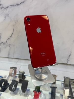 A red Apple iPhone XR with a glossy finish is displayed on a silver metal stand. The back of the phone features the silver Apple logo and 'iPhone' text.
