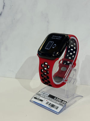 Red and black Apple Watch with a perforated sport band, displayed on a clear stand with a price tag in front, against a white marble-patterned background.