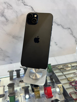 A black Apple iPhone 12 Pro displayed upright on a silver stand, with a marble-patterned background and watch displays in the foreground.