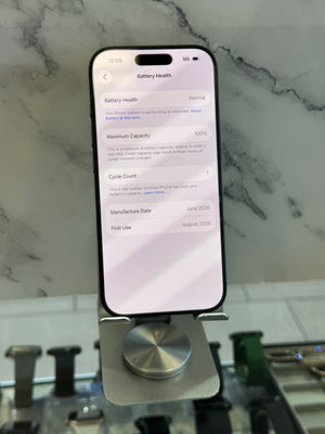 A black smartphone displayed upright on a metallic stand, showing the battery health settings screen. The phone is photographed in a real indoor setting with a marble-patterned wall and other objects visible in the background.