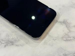 A close-up of a black smartphone lying on a white marble surface, showing the front bezel, camera opening, and glossy screen with a light reflection.