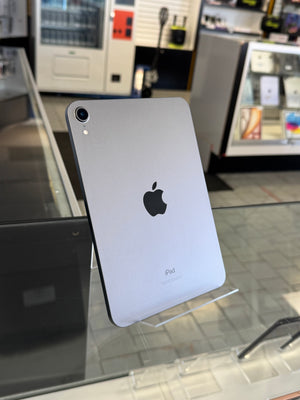 A silver Apple iPad shown from the back, resting on a clear display stand inside a retail store. The Apple logo is visible in the center.