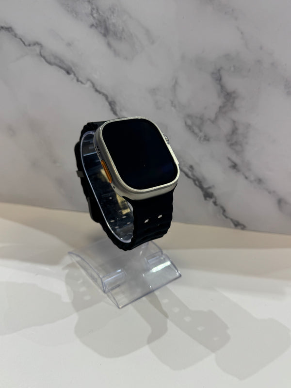 A smartwatch with a black strap and silver-toned case, displayed on a clear acrylic stand against a white marble-patterned background.