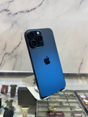 A blue Apple iPhone 12 Pro shown from the back, resting on a metal display stand in front of a marble-patterned wall.