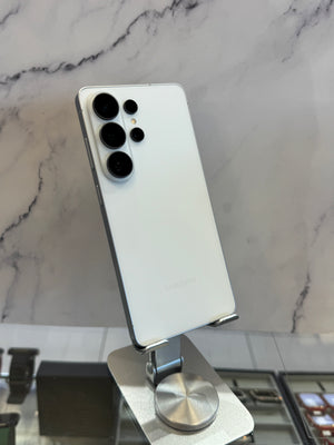 A white Samsung Galaxy smartphone shown from the back, resting on a silver metal display stand against a white marble-patterned wall.