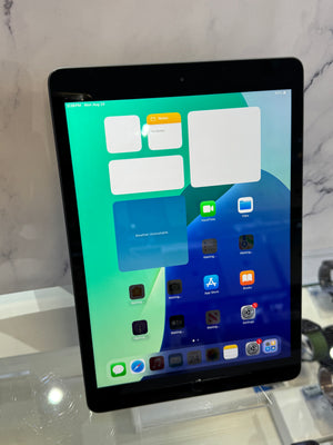 A black tablet device displaying its home screen with app icons, photographed upright on a clear display stand against a light marble-patterned background.