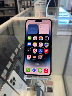 A black iPhone with a dark glass front and a black front bumper, displayed on a clear stand inside a retail store. The screen is on, showing various colorful app icons and widgets.