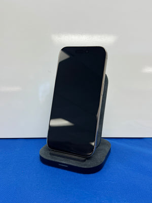 A black smartphone with a blank black screen stands upright on a dark gray charging dock or stand against a white backdrop, with a blue surface visible at the bottom.