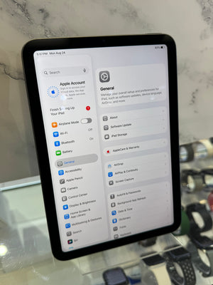 A black Apple iPad displaying the Settings app on a countertop, with a marble-patterned background and a blurred hand holding another device in the foreground.