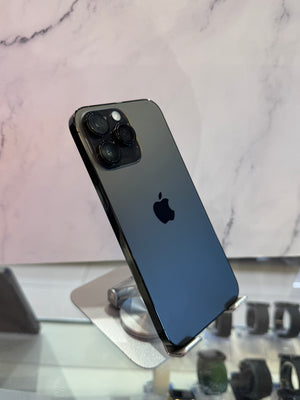 A black Apple iPhone 12 Pro shown from the back, resting on a silver metal display stand in front of a light marble-patterned background.