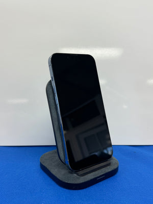 A black smartphone with a dark screen stands upright on a gray fabric display stand against a white background, with a blue surface beneath the stand.