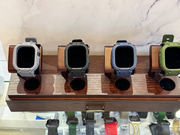 Four rugged smartwatch cases displayed on a wooden stand, shown in light gray, dark gray, muted blue, and olive green. The watches are photographed indoors against a light marble-patterned background.