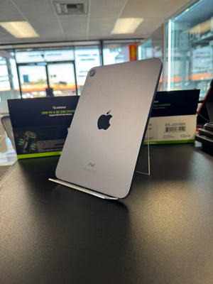 A silver Apple iPad standing upright on a clear display stand on a dark countertop, with retail packaging and a store interior visible in the background.