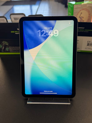A black tablet with a colorful lock screen wallpaper displayed on a clear acrylic stand on a dark tabletop, with retail boxes blurred in the background.