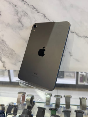 A dark gray Apple iPad shown from the back, resting on a clear display stand in front of a white marble-patterned background with watch displays visible below.