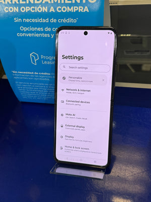 A smartphone displayed upright on a small stand, showing the Settings menu on the screen. The phone is photographed on a blue tabletop in front of a blue promotional sign with Spanish text.