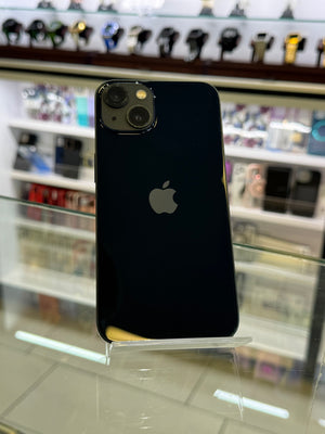 A black Apple iPhone with a glossy finish is displayed on a clear acrylic stand inside a retail store. The Apple logo is visible in the center of the back, and the background shows blurred shelves of watches and other electronics.