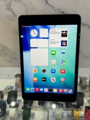 A black Apple iPad tablet standing upright on a clear display stand, showing the home screen with various app icons. The tablet is photographed in a retail or showroom setting with marble-patterned wall and other accessories blurred in the background.