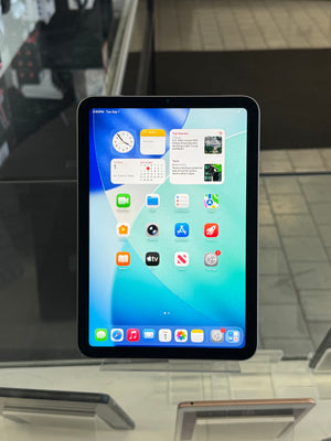 An iPad with a black bezel and colorful home screen wallpaper, displayed on a clear stand inside what appears to be a store or office.