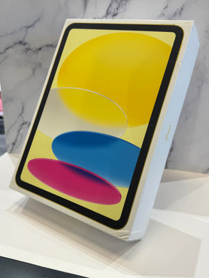 An Apple iPad in its original white retail box, standing on a white surface against a gray marble-patterned background. The screen displays a colorful abstract design with yellow, blue, and pink shapes.