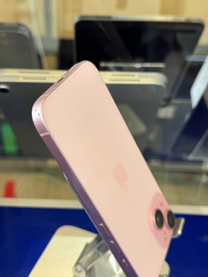 A light pink Apple iPhone shown from the back, resting on a display stand in a retail or showroom setting with other phones blurred in the background.