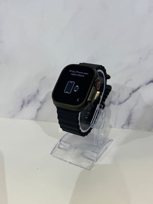 A black Apple Watch with a black band and a gold-tone side button, displayed on a clear acrylic stand against a white marble-patterned background.