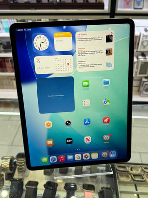An iPad with a black bezel displaying the home screen, standing on a clear display stand inside a retail store. Watches and other electronics are visible in the background.