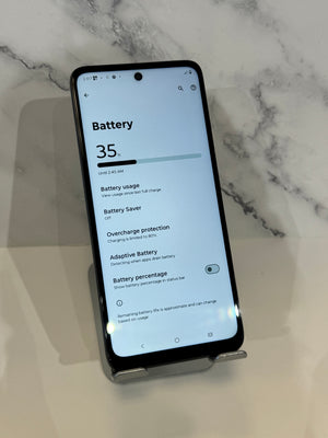 A black smartphone shown from the back, resting on a small clear stand on a light marble surface. The screen displays the battery settings page.