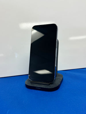 A black smartphone with a blank screen stands upright on a small dark charging dock or stand against a white wall, with a bright blue surface below.