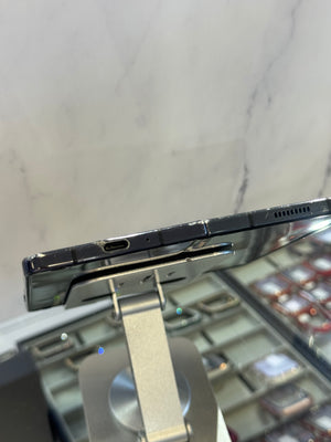 Close-up of the side edge of a black smartphone mounted on a silver display stand, showing the USB-C port and speaker grille, with a blurred retail display in the background.