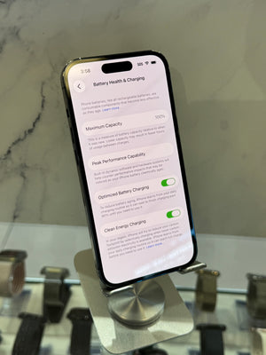 A smartphone on a metal display stand showing the Battery Health & Charging settings screen, photographed in a retail or showroom setting with marble wall and other devices blurred in the background.