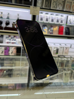 A black smartphone with a dark screen displaying the time 3:58, standing on a clear acrylic display stand inside a retail store. Shelves with various accessories are blurred in the background.