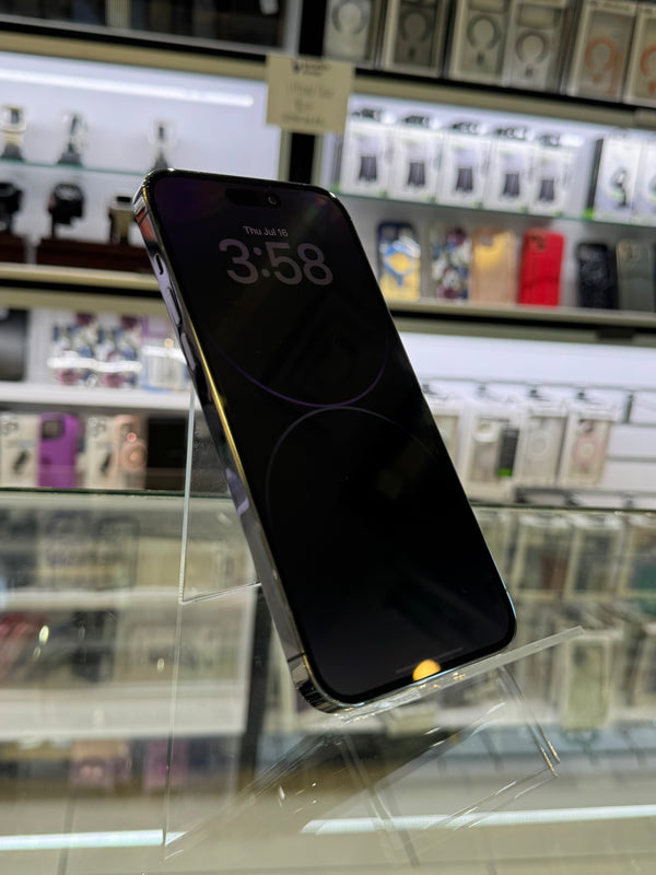 A black smartphone with a dark screen displaying the time 3:58, standing on a clear acrylic display stand inside a retail store. Shelves with various accessories are blurred in the background.