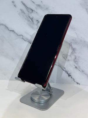 A black smartphone with a red side frame stands upright on a silver metal stand against a white marble-patterned background.
