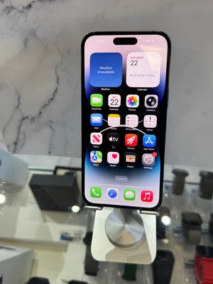 A black iPhone with its screen on, resting on a silver metal display stand. The background shows a marble-patterned wall and a blurred retail counter.