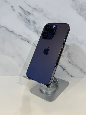 A dark blue iPhone Pro showing its back panel with the Apple logo, displayed on a silver stand against a white marble-patterned background.