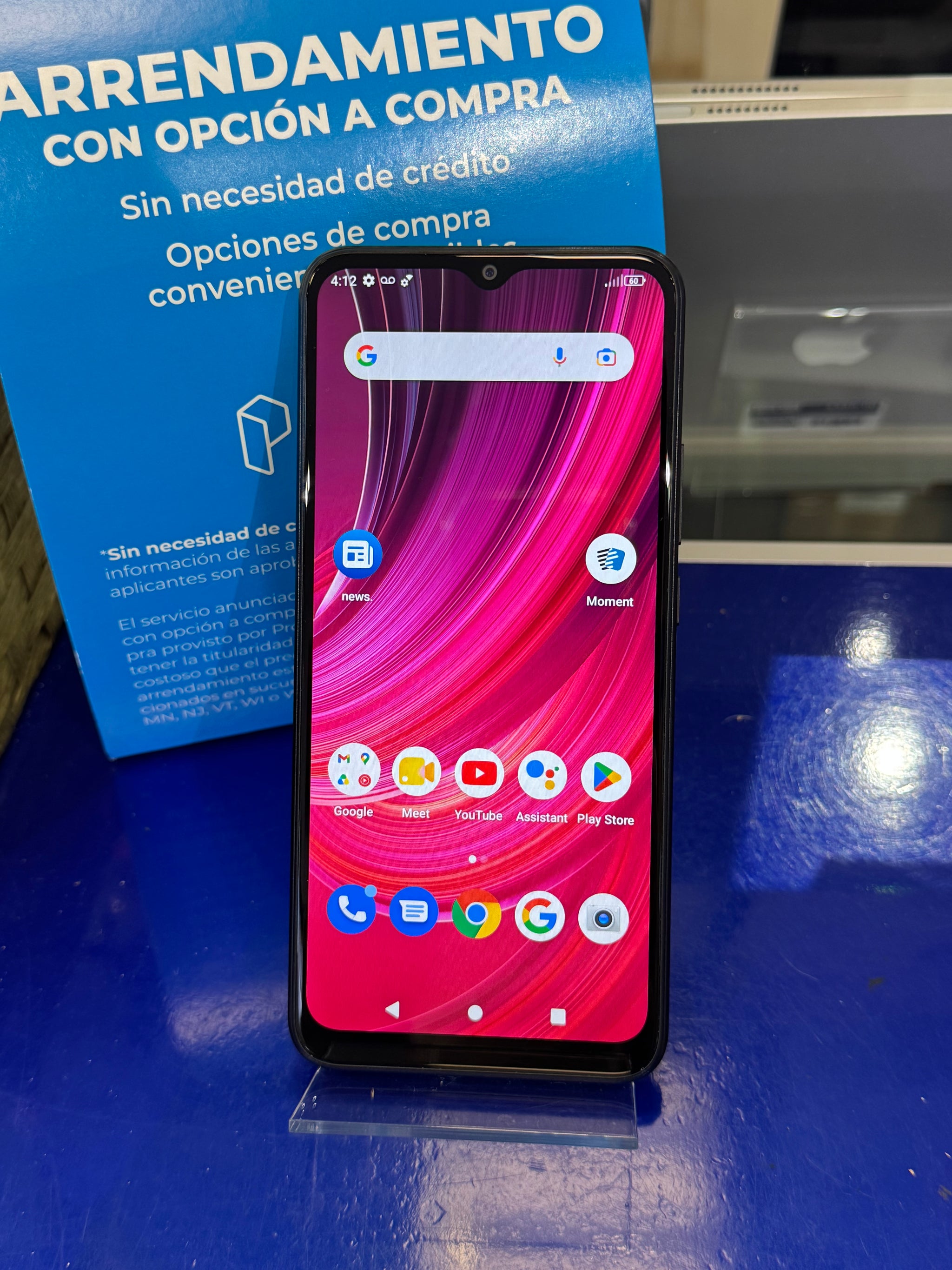 A black smartphone with a colorful pink and red wallpaper displayed on a clear stand on a blue surface, with a blue promotional sign in the background.