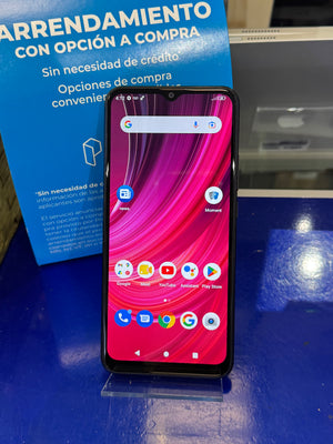 A black smartphone with a colorful pink and red wallpaper displayed on a clear stand on a blue surface, with a blue promotional sign in the background.