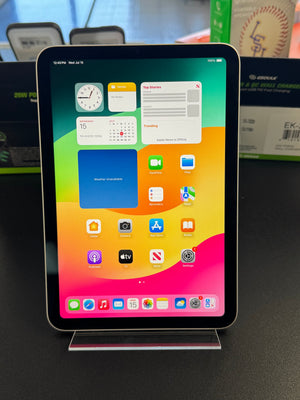 An iPad standing upright on a clear acrylic display stand, showing the home screen with colorful app icons and widgets. The device is photographed indoors on a dark tabletop with product boxes blurred in the background.