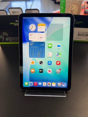An iPad with a black bezel and a colorful home screen, displayed on a clear acrylic stand on a dark tabletop.