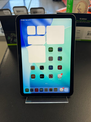 An iPad with a black bezel and colorful home screen, displayed upright on a clear acrylic stand on a dark tabletop.
