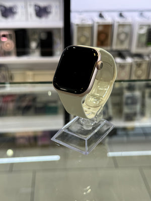A smartwatch with a cream-colored band and a black screen, mounted on a clear acrylic stand inside what appears to be a retail display.