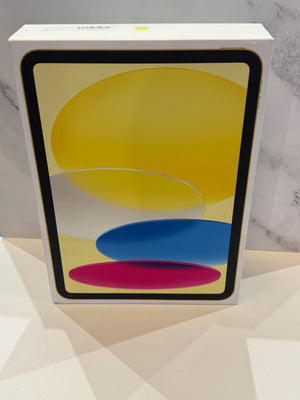 A white Apple iPad product box containing a tablet with a black bezel and a colorful screen displaying yellow, blue, and pink abstract shapes, set against a light marble and geometric wall background.