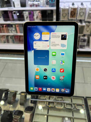 A tablet device with a black bezel and a colorful home screen, photographed in a retail or display environment with shelving visible in the background.