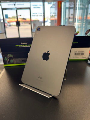 A silver Apple iPad standing upright on a clear display stand on a dark tabletop, with its retail box visible in the background near a window.