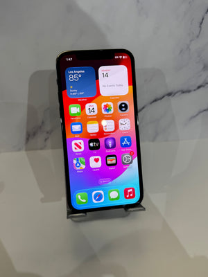 A black iPhone with its screen on, displaying the home screen, resting on a small gray stand against a white marble-patterned background.